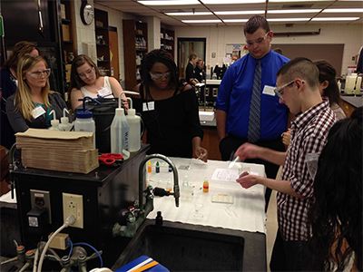 Six students wearing protective goggles, placing samples on microscope slides.