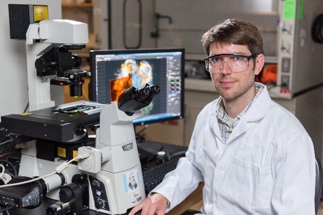 Dr. Ratcliff wearing a lab coat and safety goggles and sitting at a microscope.