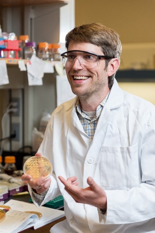 Dr. Ratcliff wearing a lab coat and safety goggles while holding a Petri dish.