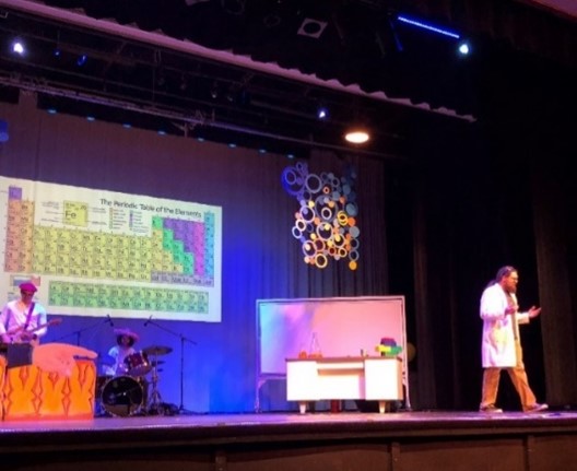 Dr. Queen stands on stage next to a man sitting at a school desk. A band stands nearby. The periodic table of elements is on the wall behind the performers.