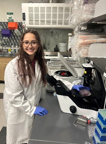 Crystal wearing a lab coat and gloves and standing in front of a microscope.