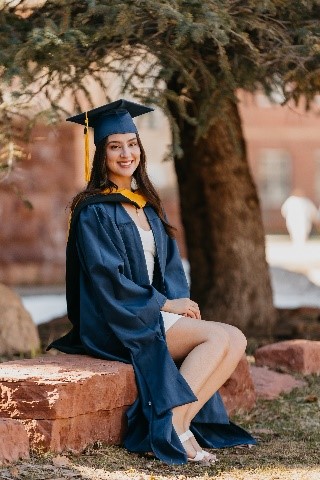 Brenda in a graduation cap and gown sitting under a tree.